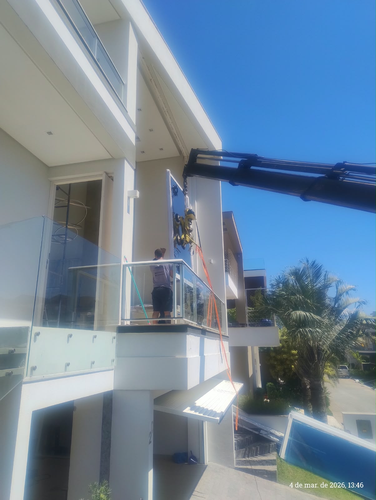 Vidro suspenso por ventosas e equipamento de içamento junto à fachada da residência, com acompanhamento da equipe na sacada.